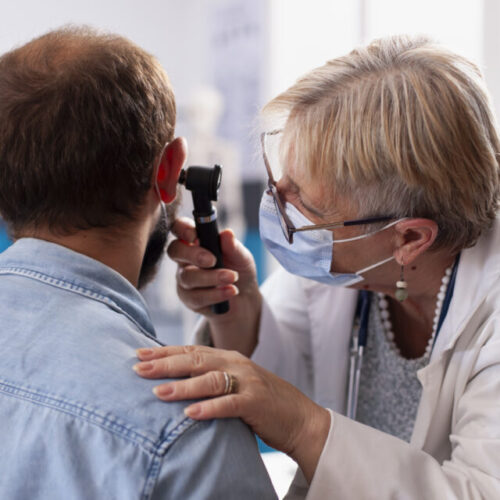 White man sits calmly in clinic room as senior ent specialist checks his ear for potential hearing issues. Old female doctor holding an otoscope during medical examination of bearded male patient.